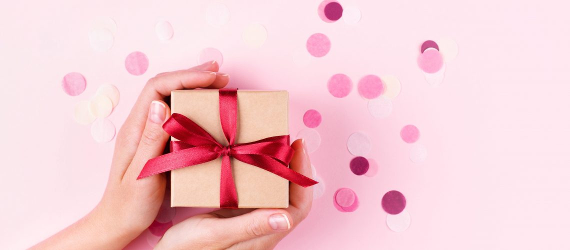 Woman's hands holding kraft gift box with red bow on pink background decorated with confetti.. Top view, holiday present concept with copy space, banner.