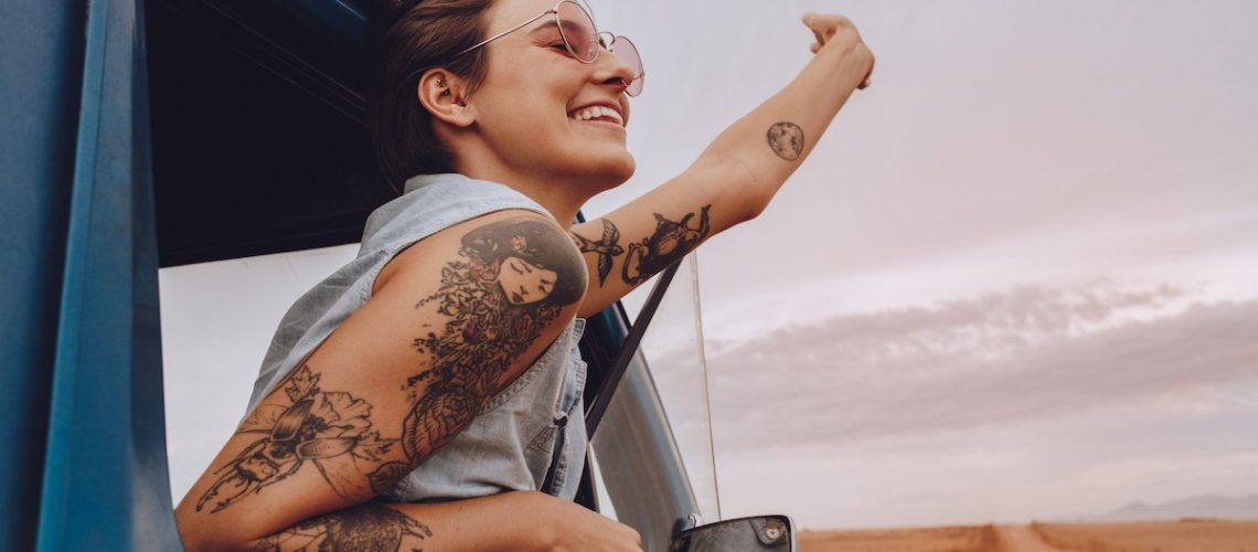 Shot of attractive young woman enjoying road trip on a summer day. Happy young female raising her hand out of the car window.